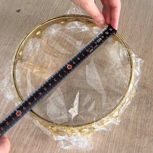Op de afbeelding: Een cirkelvormig goudkleurig metalen frame met een decoratieve geschulpte rand, verpakt in doorzichtig plastic. Een zwart meetlint is over de diameter geplaatst en toont een afmeting van ongeveer 22,2 centimeter over de opening.