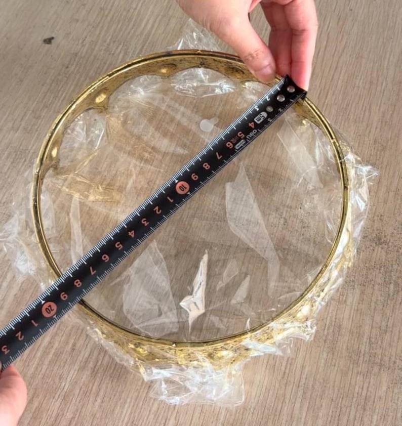 Op de afbeelding: Een cirkelvormig goudkleurig metalen frame met een decoratieve geschulpte rand, verpakt in doorzichtig plastic. Een zwart meetlint is over de diameter geplaatst en toont een afmeting van ongeveer 22,2 centimeter over de opening.