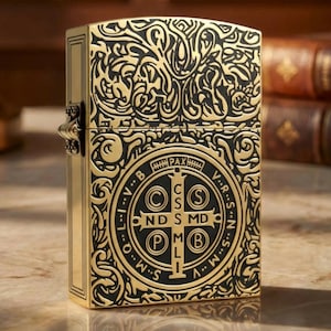 May include: A gold-colored metal lighter with intricate black engraved designs. The lighter features a detailed pattern on the front and back, with a circular emblem containing text. The lighter is rectangular and has a hinged lid.
