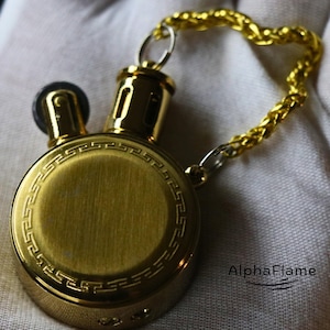 Peut inclure: Un briquet rond de couleur or avec un motif décoratif sur le bord. Il est muni d'une chaîne et d'un petit anneau. Le briquet a un dessus noir et le texte "AlphaFlame" est visible.