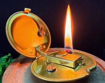Puede incluir: Un encendedor de bolsillo abierto de color dorado con una llama encendida. El encendedor es redondo con una tapa con bisagras y un puerto de combustible. La llama es alta y brillante sobre un fondo oscuro. El encendedor está sobre una superficie de madera.