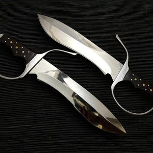 May include: Two polished steel blades with curved edges and dark wooden handles. The handles are adorned with small gold-colored studs. The blades have a curved design and a shiny, reflective surface, set against a black background.