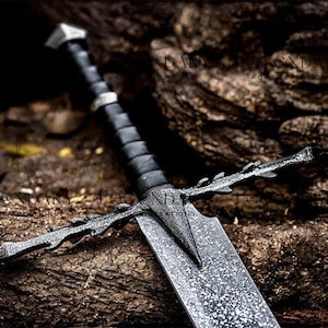 Può includere: Immagine dettagliata di una spada medievale con una lama strutturata e una guardia decorata. L'elsa è avvolta in materiale nero e il pomo ha una forma geometrica. La spada è appoggiata su una superficie marrone strutturata.