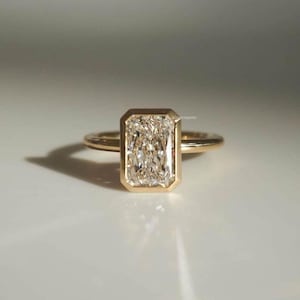Könnte beinhalten: Ein goldener Ring mit einem rechteckigen, klaren Edelstein. Der Edelstein ist in einer Lünettenfassung gefasst, und das Band ist dünn und schlicht. Der Ring wird auf einer weißen Oberfläche präsentiert.