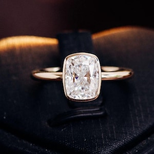 Könnte beinhalten: Ein goldener Ring mit einem einzelnen, im Kissenschliff facettierten, klaren Edelstein in einer glatten Zargenfassung. Die Ringschiene ist schmal und poliert und weist eine reflektierende metallische Oberfläche auf, die den Stein in der Mitte ergänzt.