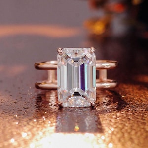 Könnte beinhalten: Ein Ring mit einem großen, klaren Edelstein im Smaragdschliff, gefasst in ein Roségoldband. Das Band besteht aus zwei parallelen, dünnen, horizontalen Metallstegen, die mit der zentralen Steinfassung verbunden sind und ein offenes Design bilden.