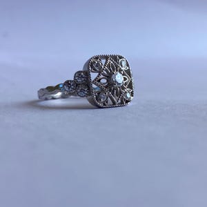 May include: A silver-toned ring featuring a square-shaped head with an intricate openwork filigree pattern. The center is set with a small round stone, surrounded by smaller stones within the filigree and three stones along the band.