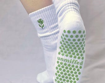 May include: White socks with green non-slip grips on the soles. The socks have a ribbed cuff and a small green embroidered leaf design. The word "unmiffed" is printed in green on the sole of the sock.