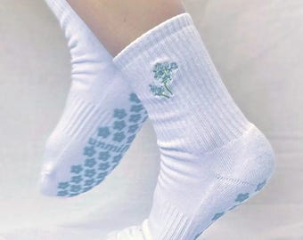 May include: White socks with a floral design embroidered near the cuff. The socks have a ribbed texture and a light blue floral pattern on the soles for grip. The image shows the socks on a person's feet.