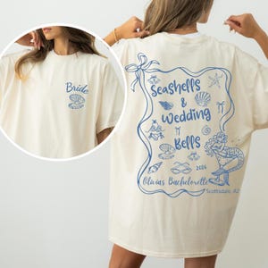 以下が含まれることがあります： クリーム色のTシャツ。前面には「Bride」の文字と貝殻、背面には「Seashells & Wedding Bells」の文字と人魚や貝殻のイラスト、そして「Olivia's Bachelorette 2026 Scottsdale, AZ」の文字が青色でプリントされています。