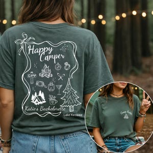 May include: A dark green t-shirt with a white graphic that says "Happy Camper" with camping-themed illustrations. The text "Katie's Bachelorette" and "Lake Keowee" are also included. A smaller version of the design is on the front.
