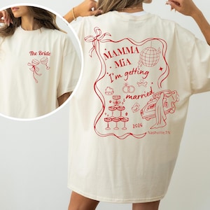 Puede incluir: Camiseta color crema con gráficos rojos. La parte delantera presenta el texto "The Bride" con copas y lazos. La parte trasera muestra "MAMMA MIA I'm getting married" con ilustraciones de boda, el año 2026 y "Nashville, TN".