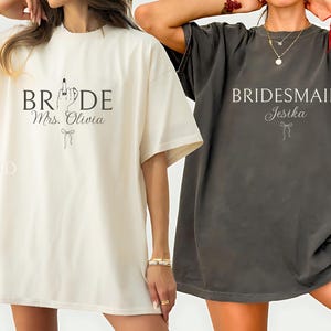 May include: Two oversized t-shirts, one cream and one charcoal grey. The cream shirt says "BRIDE Mrs. Olivia" with a bow graphic. The grey shirt says "BRIDESMAID Jesika" with a bow graphic. Both shirts are short-sleeved.