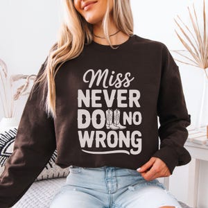 May include: A dark brown sweatshirt featuring the white text "Miss Never Do No Wrong" and a pair of cowboy boots. The sweatshirt has a crew neck and long sleeves.