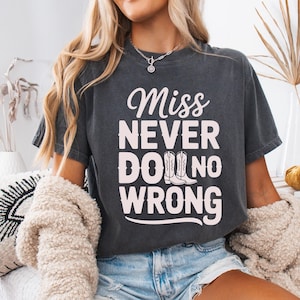 Op de afbeelding: Donkergrijs t-shirt met de witte tekst "Miss Never Do No Wrong", met een paar cowboylaarzen. Het shirt heeft een vintage, versleten uitstraling.