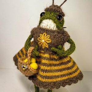 May include: A whimsical, handmade knitted frog figurine wearing a brown and yellow striped dress with a yellow flower detail. The frog holds a small basket with yellow objects and has antennae. The frog is a unique, handcrafted item.