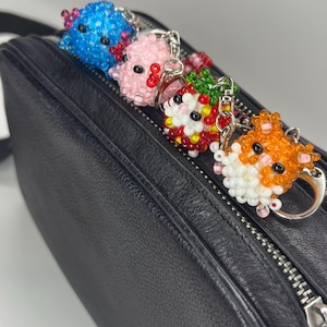 May include: Four beaded animal keychains in blue, pink, red, and orange, each with a silver keyring. The keychains are placed on a black leather bag. The keychains are in the shape of small animals.