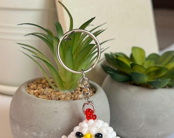 May include: A beaded chicken keychain with a silver keyring. The chicken is white with red, black, and yellow accents. The word "GOALS" is visible on a notebook in the background. Small potted plants are also in the image.