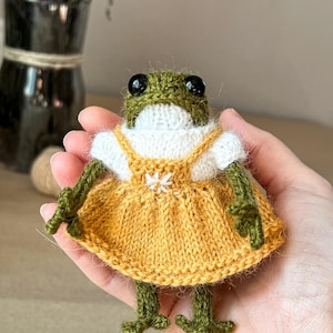 May include: A small, hand-knitted green frog figurine with large black bead eyes. The frog wears a white short-sleeved top under a mustard yellow pinafore dress featuring a small white embroidered flower detail on the front chest area.