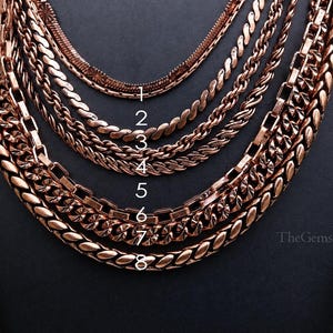 Puede incluir: Una colección de ocho cadenas de metal en tonos cobre dispuestas en capas. Cada cadena presenta un estilo de eslabón distinto, numerado del uno al ocho en texto blanco. Las cadenas varían en grosor y textura, mostrando un acabado metálico.