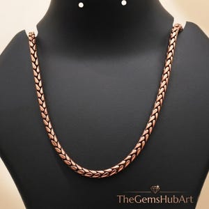 May include: A copper-toned chain necklace featuring a repeating woven wheat or foxtail link pattern. The metallic links have a warm, reddish-brown hue with a polished finish. The text TheGemsHubArt is printed in white at the bottom right corner.