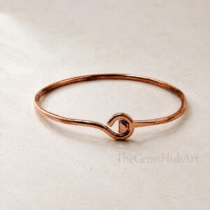 Puede incluir: Una pulsera rígida de metal color cobre con un diseño de alambre circular liso que se curva hacia un cierre de estilo gancho en la parte delantera. La superficie de la pulsera tiene un acabado metálico cálido con sutiles variaciones de tono.