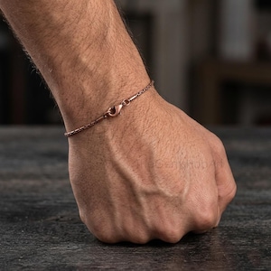 May include: A thin copper-toned chain bracelet featuring a small lobster claw clasp. The bracelet is composed of delicate, uniform rectangular links that wrap around the wrist, showcasing a metallic finish with a warm, reddish-brown hue.