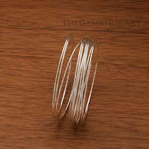 May include: A silver-colored spiral bangle bracelet consisting of multiple thin, continuous wire loops that overlap to create a layered, coiled appearance. The metallic surface reflects light, highlighting the smooth, rounded texture of the wire.