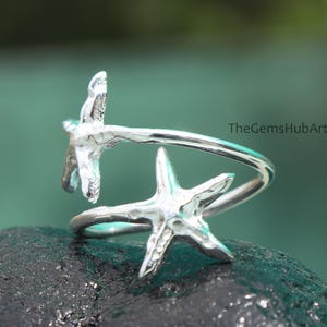 Puede incluir: Un anillo plateado con dos diseños de estrellas de mar. El anillo tiene un diseño envolvente con las estrellas de mar mirando en direcciones opuestas. El anillo está sobre una superficie oscura y texturizada con un fondo verde borroso.