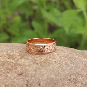 Può includere: Un anello a fascia color oro rosa caratterizzato da un motivo floreale e a tralci in rilievo sulla superficie esterna. L'anello in metallo ha una finitura lucida e riporta la scritta TheGemsHubArt in piccole lettere nere vicino al bordo.