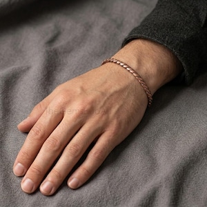 Pode incluir: Uma pulseira de cor cobre metálico com um design trançado semelhante a uma corda. A pulseira é usada no pulso e apresenta uma superfície polida e reflexiva com um padrão em espiral contínuo que envolve toda a circunferência.