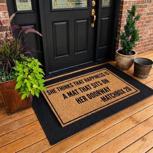 May include: A rectangular tan coir doormat with a black border and black text that reads: SHE THINKS THAT HAPPINESS IS A MAT THAT SITS ON HER DOORWAY -MATCHBOX 20. The mat is layered on top of a larger solid black rectangular mat on a wooden porch.