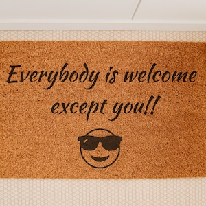 May include: A rectangular doormat made of natural coir fiber. The mat features the phrase "Everybody is welcome except you!!" in black script, along with a smiling face wearing sunglasses. A humorous welcome mat.