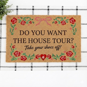 Op de afbeelding: Een rechthoekige kokosmat met de tekst "DO YOU WANT THE HOUSE TOUR? Take your shoes off!" in bruin. De mat is versierd met rode rozen en groene bladeren, en een roze strik.