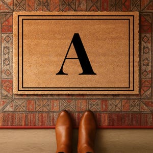 Op de afbeelding: Een rechthoekige deurmat met een zwarte rand en een grote zwarte letter "A" in het midden. De mat heeft een natuurlijke bruine kleur en ligt op een patroonkleed. Bruine leren schoenen zijn onderaan zichtbaar.