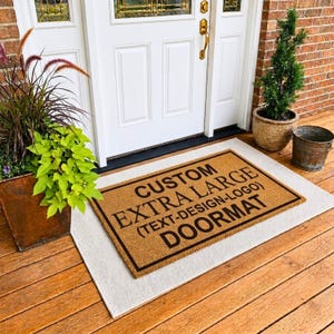 Op de afbeelding: Een rechthoekige, beige kokosmat met een zwarte rand en zwarte tekst die luidt CUSTOM EXTRA LARGE (TEXT-DESIGN-LOGO) DOORMAT. De mat ligt op een lichtgekleurd kleed op een houten veranda voor een witte deur en een bakstenen muur.