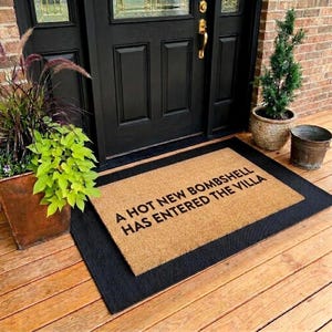 May include: A rectangular doormat with the text "A HOT NEW BOMBSHELL HAS ENTERED THE VILLA" in black letters. The mat is tan with a black border, placed on a wooden porch in front of a black door.