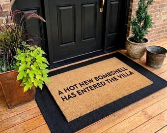 May include: A rectangular doormat with the text "A HOT NEW BOMBSHELL HAS ENTERED THE VILLA" in black letters. The mat is tan with a black border, placed on a wooden porch in front of a black door.