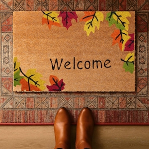 Op de afbeelding: Een rechthoekige deurmat met het woord "Welcome" in zwarte letters. De mat is bruin met kleurrijke herfstbladeren in de hoeken. Bruine laarzen zijn zichtbaar aan de onderkant van de afbeelding, staand op een patroonkleed.