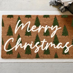 May include: A rectangular doormat with a brown background and a repeating pattern of green Christmas trees. The words "Merry Christmas" are written in white script across the center. The mat is bordered by a white fringe.