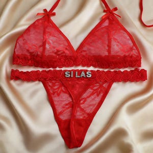 May include: Red lace lingerie set featuring a triangle bralette with bow accents and a matching thong. The thong has the name "SILAS" spelled out in silver letters. The set is displayed on a satin surface.