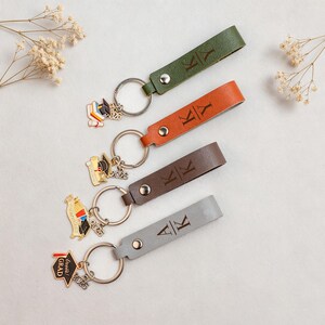 Op de afbeelding: Vier leren sleutelhangers in groen, oranje, bruin en grijs. Elke sleutelhanger heeft een metalen ring en een diploma-thema bedel met het jaar 2026. De leren bandjes zijn voorzien van initialen.
