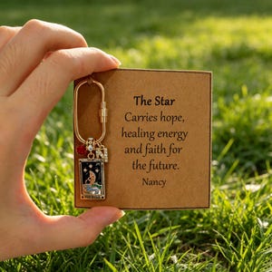 May include: A gold-toned oval carabiner keychain featuring a rectangular tarot card charm depicting a star, a small red crystal bead, and a letter N charm. The keychain is attached to a square brown card with the text The Star Carries hope, healing energy and faith for the future. Nancy.