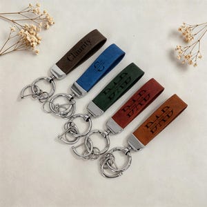 May include: Five leather keychains in brown, blue, green, red, and tan. Each keychain has a silver-colored metal clasp and a leather strap with engraved text. The text includes names and the word "DAD".