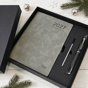 May include: A grey 2027 schedule notebook with a textured cover featuring the name Izzy, presented in a black gift box alongside a black pen and a metal ink refill. The notebook cover displays the text 2027 Schedule Notebook in black font.
