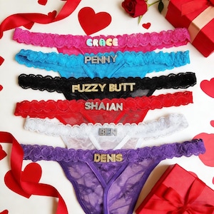 May include: A collection of six lace thong underwear in various colors, including pink, blue, black, red, white, and purple. Each thong features a name or phrase spelled out in decorative letters. Valentine's Day theme with hearts and a red rose.