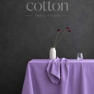 May include: A solid lavender tablecloth draped over a rectangular table. On the table sits a white ribbed vase containing three purple allium flowers and a clear glass tumbler. The text "cotton TABLE STUDIO" is visible on the dark gray wall.