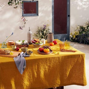May include: A mustard yellow linen tablecloth draped over a wooden table, set with ceramic plates, bowls, a gray pitcher, a bottle, glassware, a blue linen napkin, a wooden cutting board with sliced bread, and fresh tomatoes and fruit.