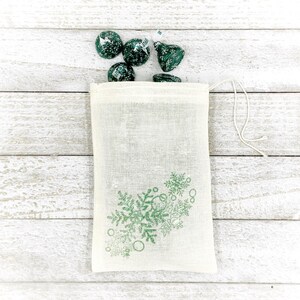 K&ouml;nnte beinhalten: Eine kleine, wei&szlig;e, Kordelzugtasche mit einem gr&uuml;nen Schneeflockenmuster. Die Tasche ist mit gr&uuml;nen und wei&szlig;en Schokoladenbonbons gef&uuml;llt.