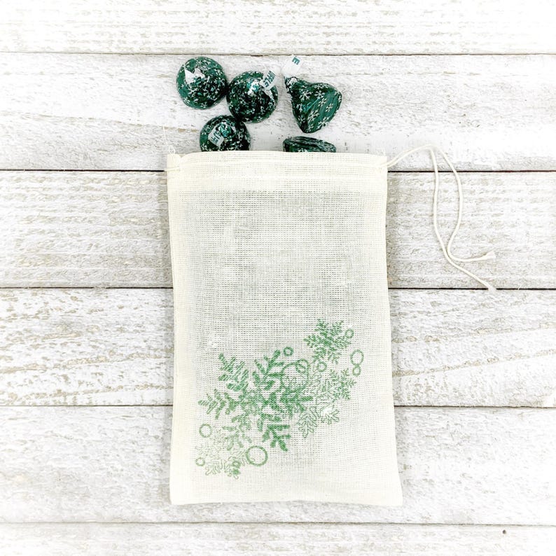 K&ouml;nnte beinhalten: Eine kleine, wei&szlig;e, Kordelzugtasche mit einem gr&uuml;nen Schneeflockenmuster. Die Tasche ist mit gr&uuml;nen und wei&szlig;en Schokoladenbonbons gef&uuml;llt.